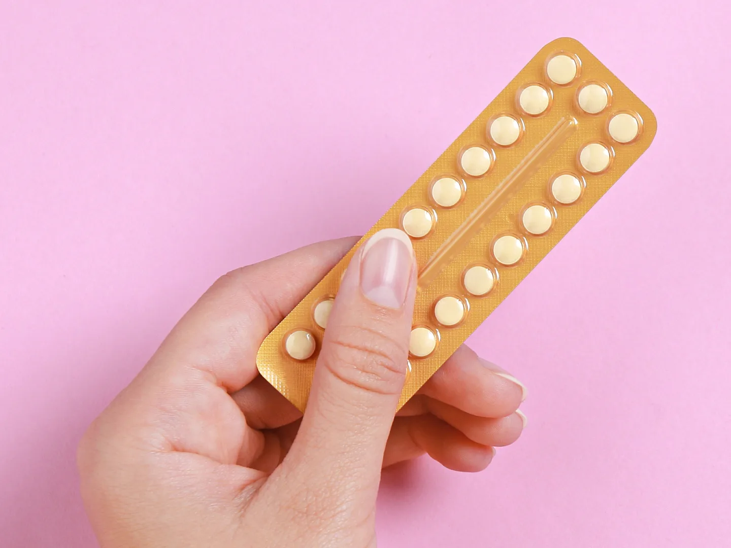 A hand holding a full blister pack of tablets on a pink background